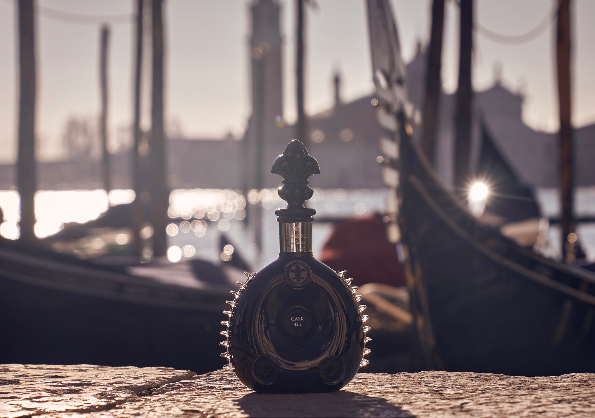 Louis III Rare Cask 42.1 cognac black in a Baccarat decanter on side of dock in Venice with boats in the background